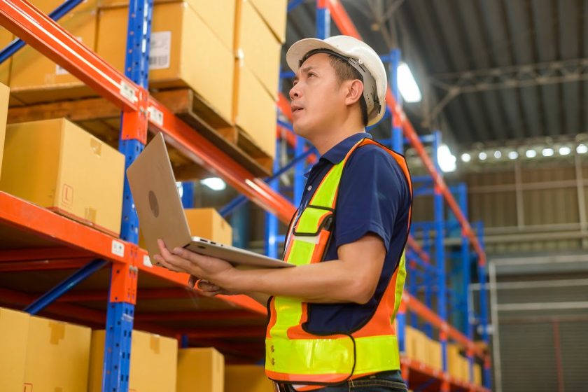 Young asian male worker wearing helmet using laptop in modern warehouse.