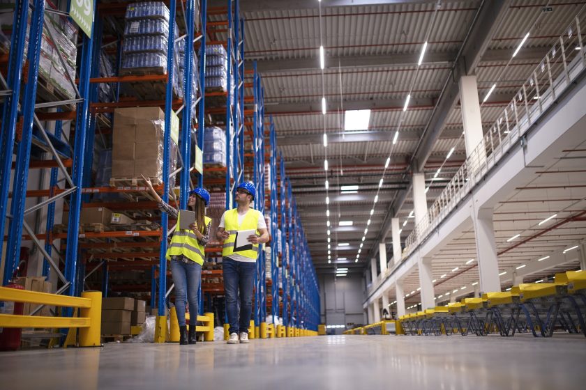 Shot of two workers walking through large warehouse center, observing racks with goods and planing distribution to the market.